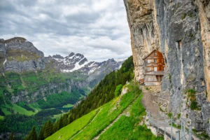 A wooden building beside a steep cliff above a green mountain valley in Switzerland.